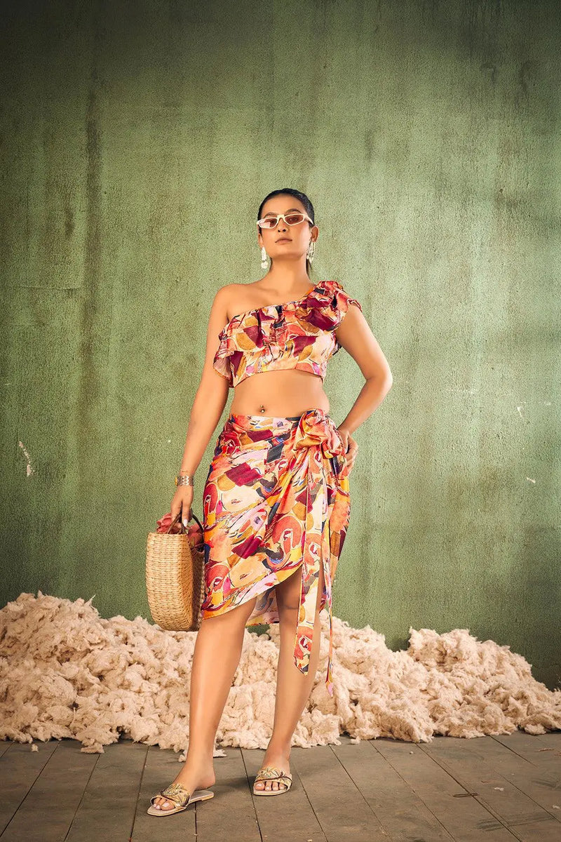 Woman in a colorful one-shoulder top and floral skirt standing against a textured green wall with cotton bales. [Abstract Print Beach Wear - Plus Size - Women]